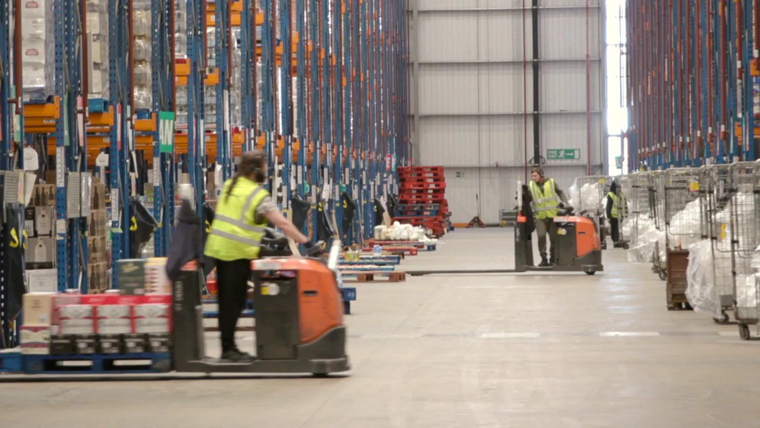 Wide-angle view of a high-bay warehouse with workers operating electric pallet trucks along a central aisle, flanked by tall racking and staged pallets. Ideal for illustrating independent supply chain integrity audits, goods receiving verification, pick accuracy checks, and logistics performance assurance in regulated retail environments.