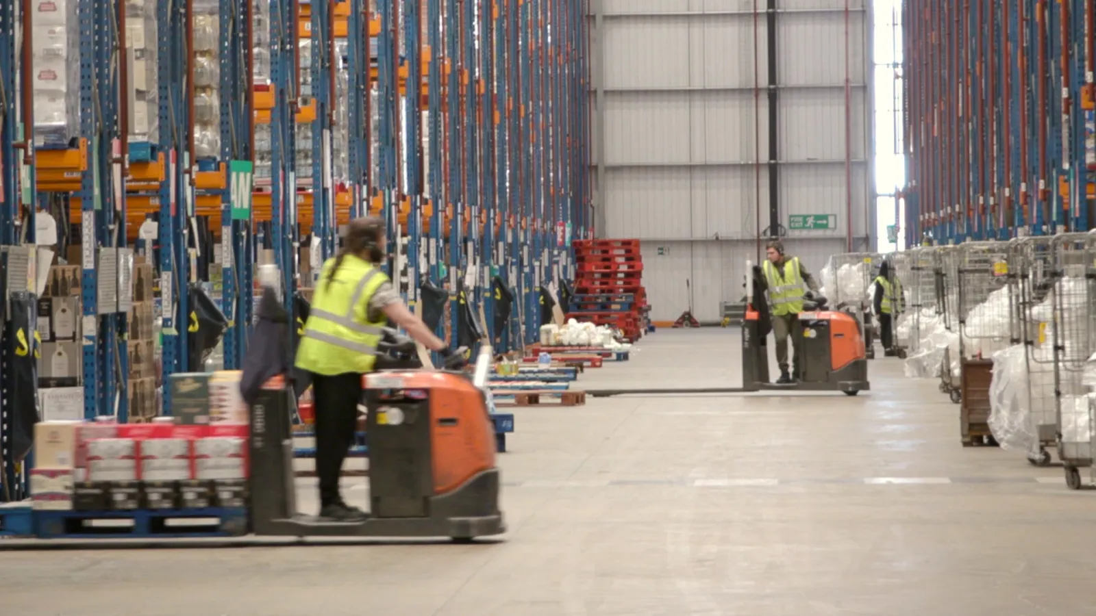 Wide-angle view of a high-bay warehouse with workers operating electric pallet trucks along a central aisle, flanked by tall racking and staged pallets. Ideal for illustrating independent supply chain integrity audits, goods receiving verification, pick accuracy checks, and logistics performance assurance in regulated retail environments.